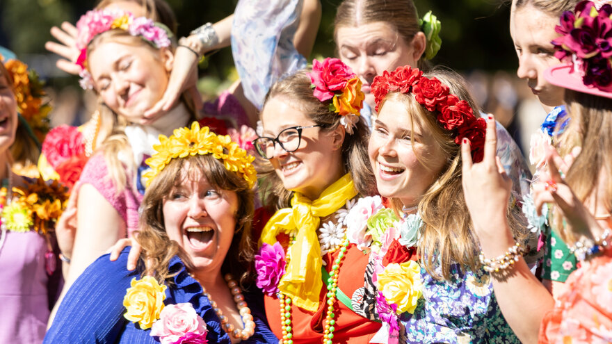 Glade studenter fra pikekoret Ivar under immatrikuleringsseremonien. De er kledd i sterke farger, og med blomsterkranser i håret og rundt halsen. 