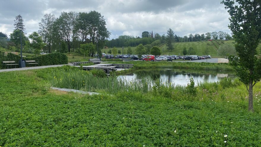 Tett på ser vi endelig den lille dammen, men vi ser også en stor parkeringsplass. Eplehagen og Ås kirke utgjør bakgrunnen. 