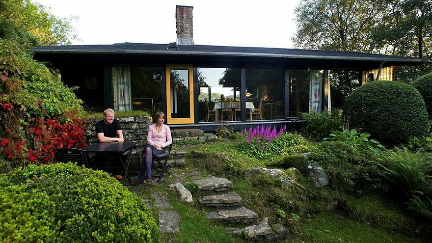 Hage designet av Torborg Zimmer med sort funkishus i bakgrunnen. To personer sitter ved et bord foran huset.